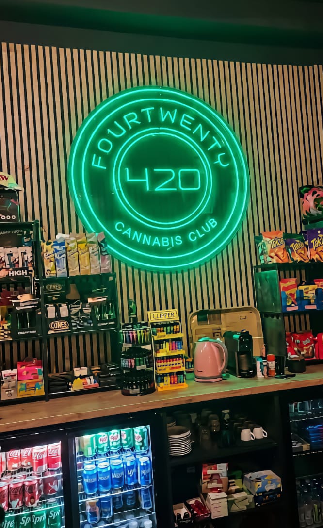 Enseigne lumineuse en n&eacute;on vert avec le logo du club de cannabis Four Twenty &agrave; Valence, install&eacute;e au-dessus d'un comptoir de bar rempli de snacks, de boissons et de briquets Clipper.