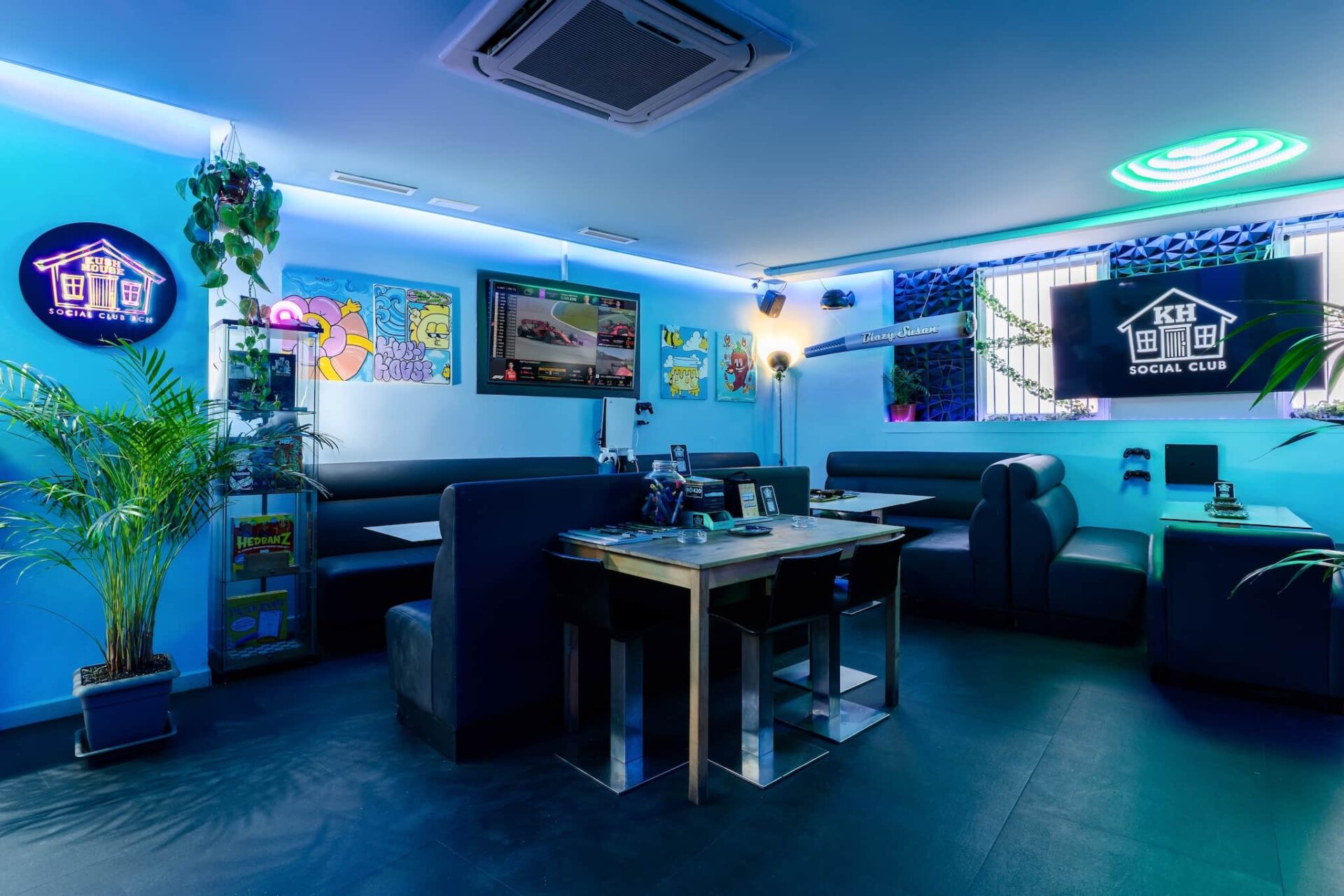 Int&eacute;rieur du Kush House Social Club &agrave; Barcelone, avec &eacute;clairage ambiant bleu, banquettes, &eacute;crans de jeux, &oelig;uvres murales color&eacute;es et plantes en pot