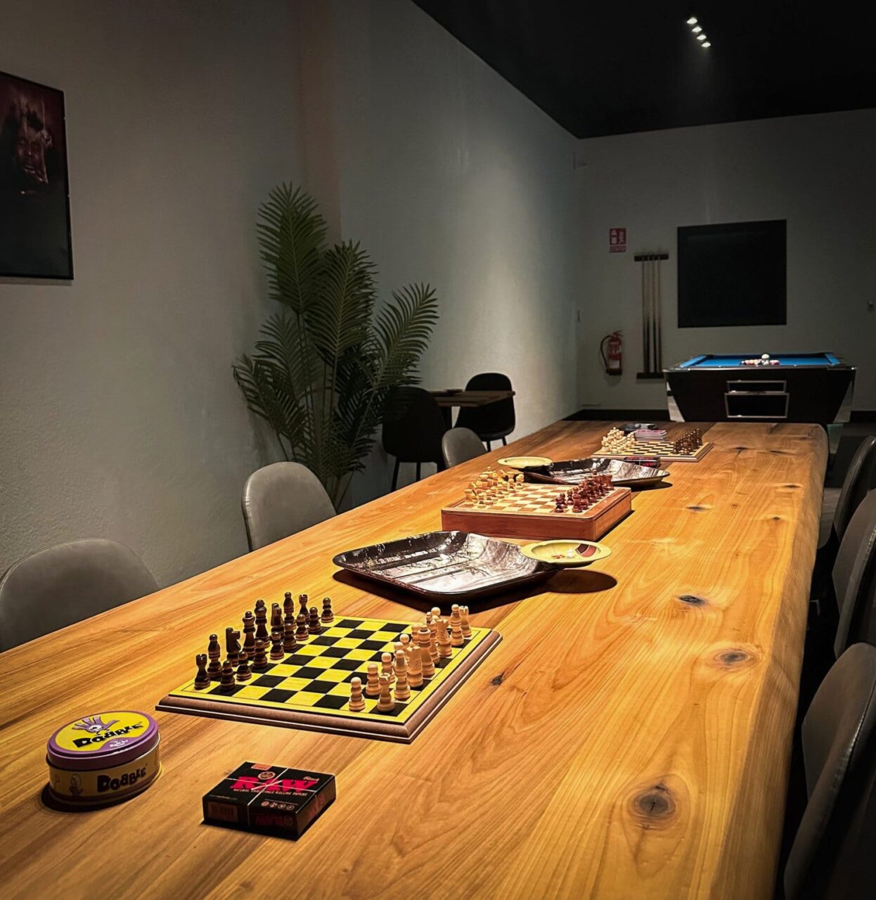 Une longue table en bois avec des &eacute;chiquiers, des jeux de soci&eacute;t&eacute; et des accessoires pour fumer au club de cannabis Four Twenty &agrave; Valence, avec un billard visible &agrave; l'arri&egrave;re-plan.