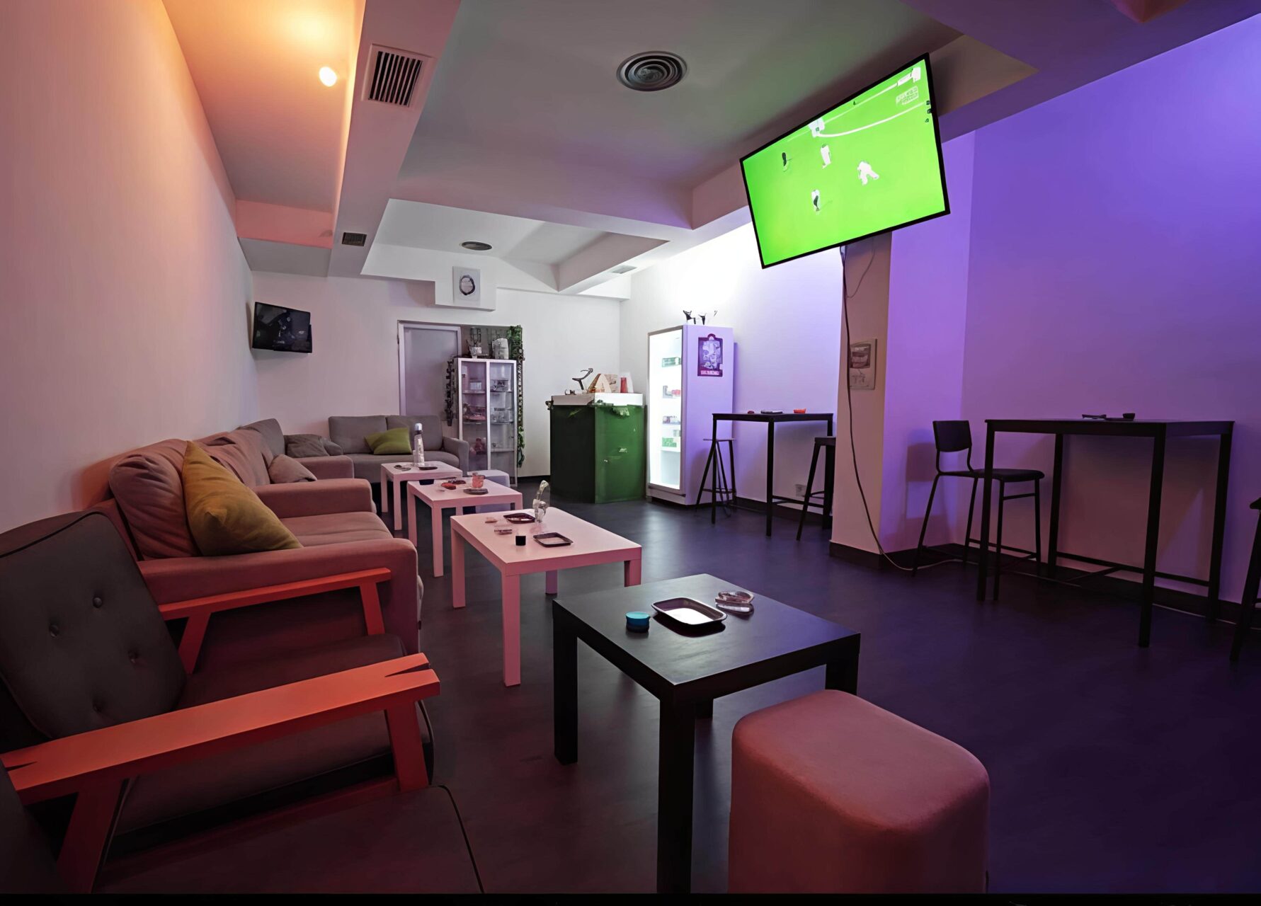 Espace lounge au Valhalla cannabis club &agrave; Madrid avec si&egrave;ges, tables hautes, un r&eacute;frig&eacute;rateur et une t&eacute;l&eacute;vision diffusant un match de football.