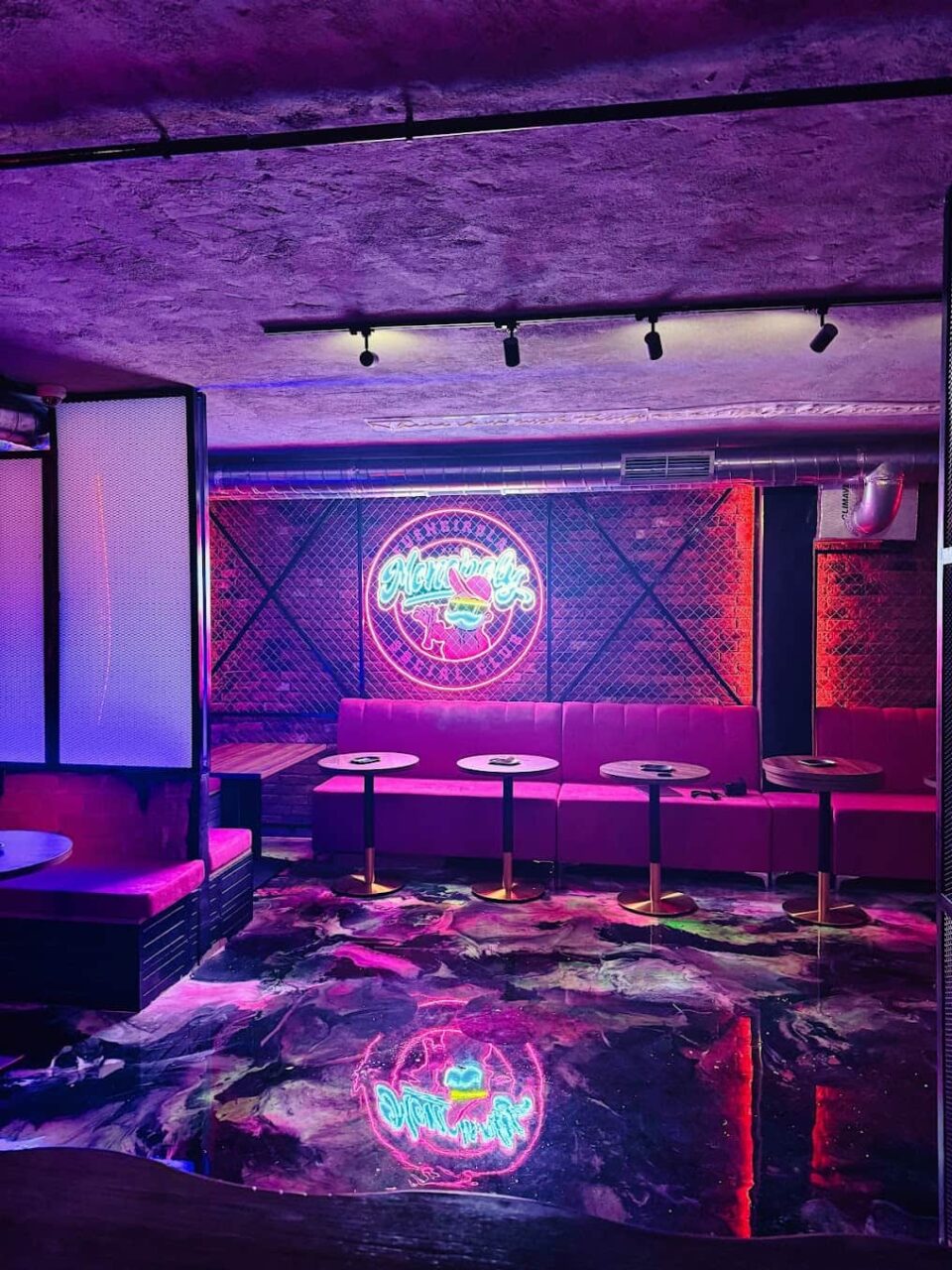 Espace lounge du Monopoly Social Club &agrave; Malaga, avec &eacute;clairage ambiant violet, mur en brique, canap&eacute;s roses capitonn&eacute;s et sol r&eacute;fl&eacute;chissant.